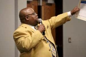 Professional man in yellow suit presenting with microphone and document at a conference.