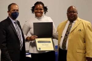 A woman receiving an award or certificate with two men in formal attire, at a professional event.