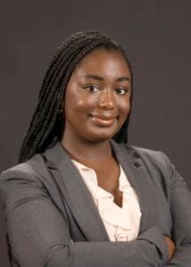 A woman in professional attire with braided hair, smiling confidently.