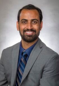 A man in a gray suit and blue shirt smiling for a professional headshot.