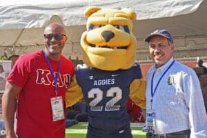 Two men and a mascot at a university event, smiling outdoors in sunny weather.