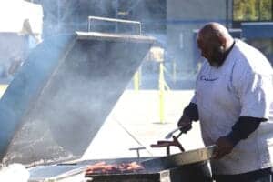 Grilling outdoors with a man cooking meat on a barbecue grill at a sunny event.