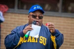 Blue Phi Beta Sigma cap and sunglasses worn by a man speaking into a microphone at an outdoor event.