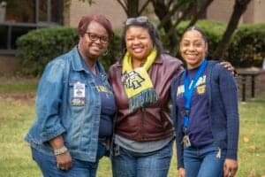 A woman and two young women smiling outdoors at a community event.