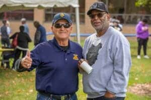 Two men smiling outdoors, one giving a thumbs-up, at a community event.