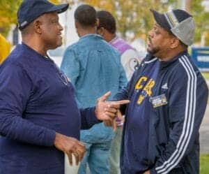 Two men having a conversation at an outdoor event, wearing casual and team apparel.