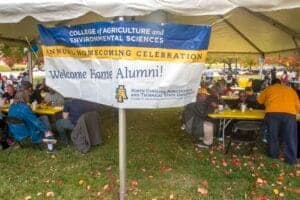 Celebration banner at college homecoming event with alumni gathering outdoors.