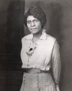 A woman in vintage attire with glasses, black hair, and a confident expression, posed for a formal p.