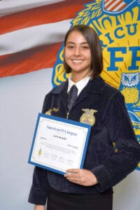 Graduation woman holding FFA degree certificate at ceremony with FFA emblem background.