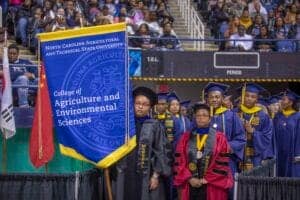 Graduates in caps and gowns at college commencement ceremony with university banner in background.