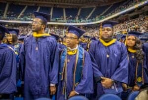 Graduates in caps and gowns at a university graduation ceremony in a large stadium.