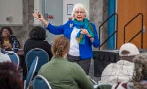 A woman speaking at a conference with an audience, holding a microphone and gesturing with her hand.