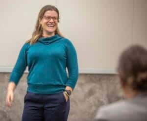 A woman smiling and speaking in a classroom or seminar setting.