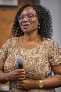 Empowered woman speaking at a conference, holding a microphone, wearing glasses and a gold lace dres.
