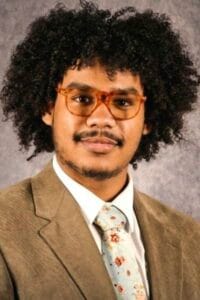 A young man with curly hair wearing glasses and a suit, smiling confidently.