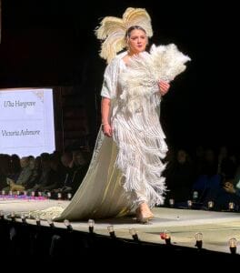Elegant fashion model walking runway in elaborate white feathered gown at a fashion show.