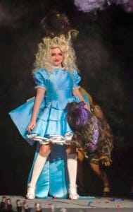 A woman dressed as Alice from Alice in Wonderland holding a small dog on stage.