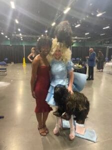 Two women in costumes with a dog at an indoor event, smiling and posing for photos.