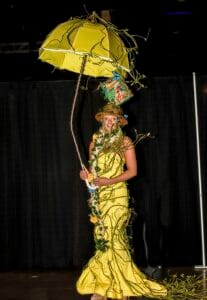 A woman dressed in a yellow gown holding a yellow umbrella decorated with vines and leaves.