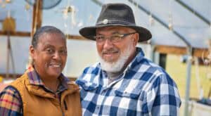 A woman and a man smiling outdoors, wearing casual clothing and hats, in a greenhouse or agricultura.