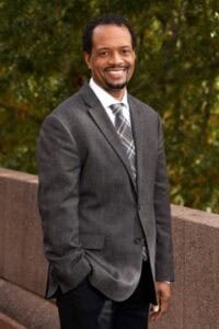 A man in a gray suit smiling outdoors with autumn foliage in the background.