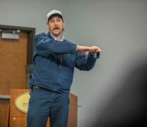 Man giving a presentation or speech in an indoor setting with a gray background.