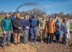 Group of diverse professionals and military personnel at outdoor event with large metal arch in back.