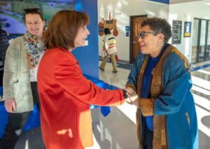 A woman in a red blazer greeting an older woman with glasses at a conference or event.