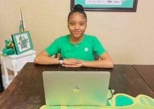 A young girl sitting at a desk with a laptop, smiling, in a classroom or office setting.