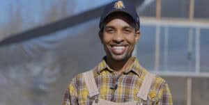 A smiling man wearing a yellow plaid shirt and a navy cap with an "A" logo, standing outdoors in fro.