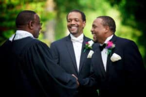 Three men in wedding attire smiling and shaking hands outdoors during wedding ceremony.
