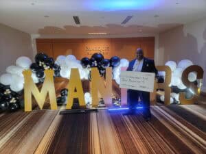 Man holding oversized check at corporate event with large "MANES" letters and balloon decorations.