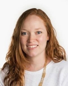 A smiling woman with long red hair wearing a white top and gold necklace.