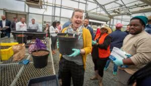 Bright woman holding a plant pot in a greenhouse with diverse people engaging in gardening activitie.