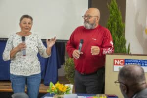 Two speakers presenting at a community event with a projector screen and greenery in the background.