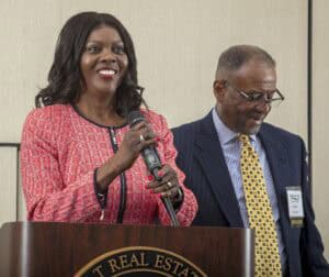 Empowered woman speaking at a conference podium with a man beside her, both in professional attire.