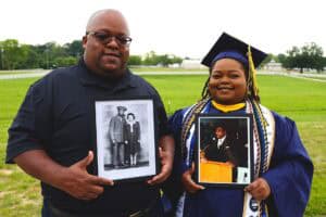 Graduation celebration featuring a young woman in cap and gown holding a framed photo, standing outd.