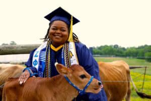 Happy graduate in cap and gown with calf on farm, celebrating academic achievement and rural life.