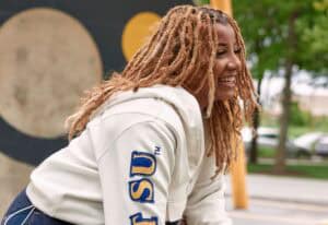 A woman with dreadlocks smiling outdoors, wearing a white hoodie with "SU" logo.