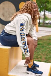 Young woman with dreadlocks sitting outdoors on a yellow bench, smiling and wearing casual sportswea.