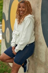 Bright smiling woman with curly hair in casual hoodie and denim shorts, leaning against graffiti wal.