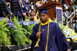 Graduation student in cap and gown at ceremony with audience and floral decorations.