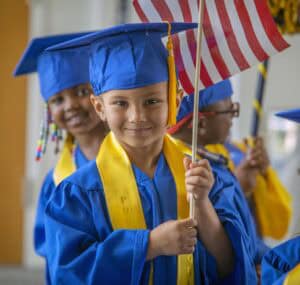Graduating children in caps and gowns holding American flags at a school ceremony.