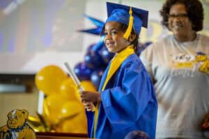 Graduating student in blue cap and gown holding diploma at school ceremony.