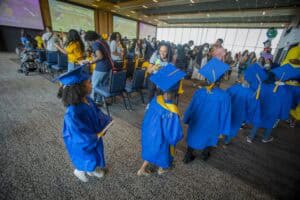 Graduates in blue caps and gowns at a graduation ceremony in a large indoor venue.