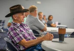 Elderly man in a plaid shirt and cowboy hat attending a conference or seminar.