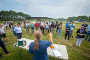 Community outdoor event with a speaker addressing a diverse group of people in a green field.