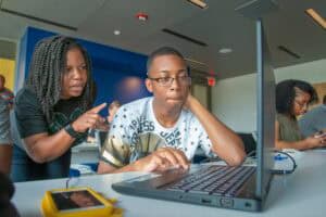 A woman instructing a young man working on a laptop in a modern classroom setting.