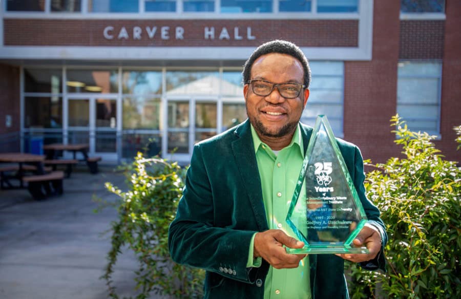 Celebrating 25 Years: Man holding an award outside Carver Hall, smiling for the camera.