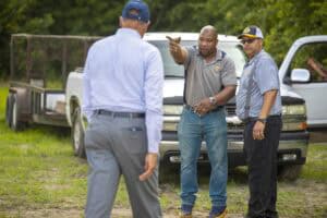 A man in a gray shirt and jeans points and discusses with two other men outdoors near a white pickup.